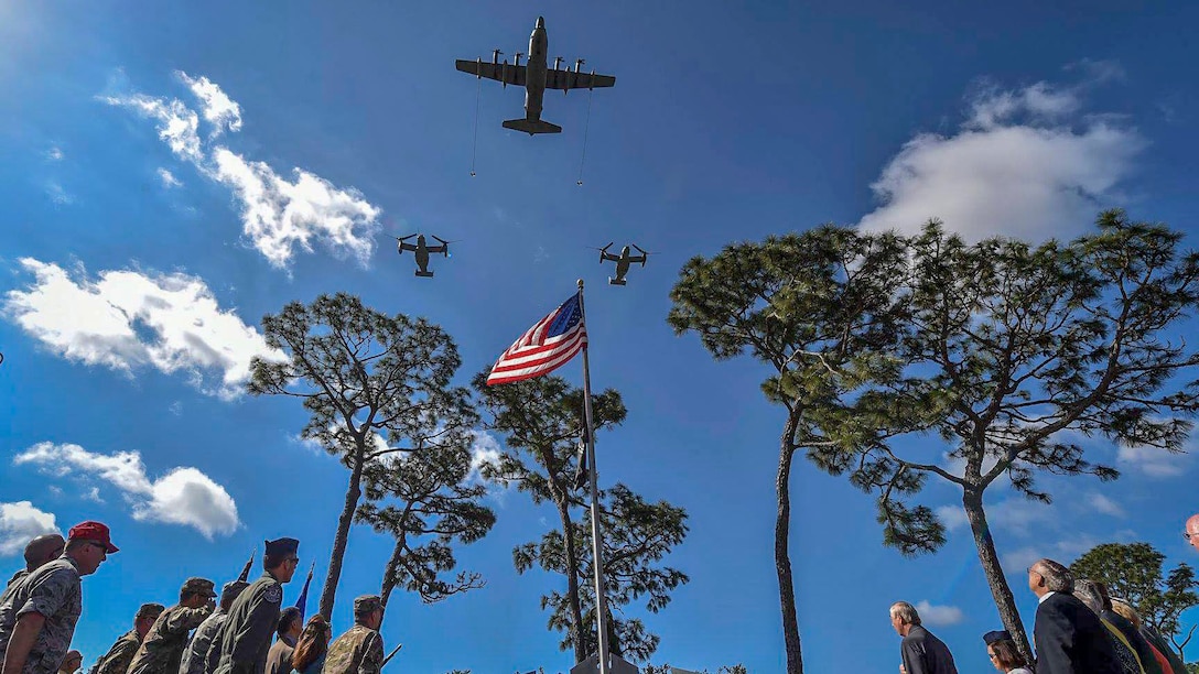 Spectators around a U.S. flag look up as three aircraft fly in formation in blue skies.