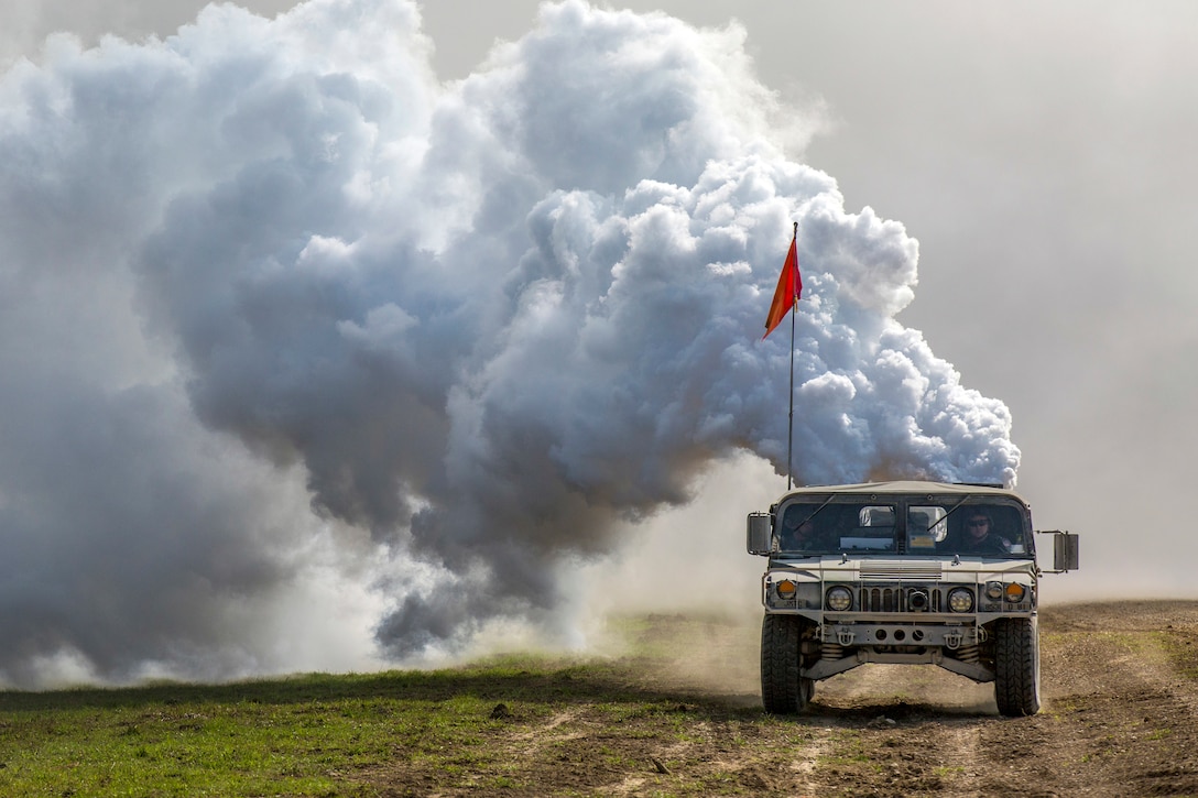 Smoke billows out behind a Humvee with a red flag displayed driving on a dirt road.