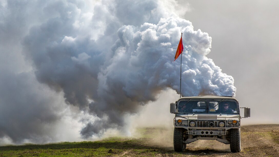 Smoke billows out behind a Humvee with a red flag displayed driving on a dirt road.