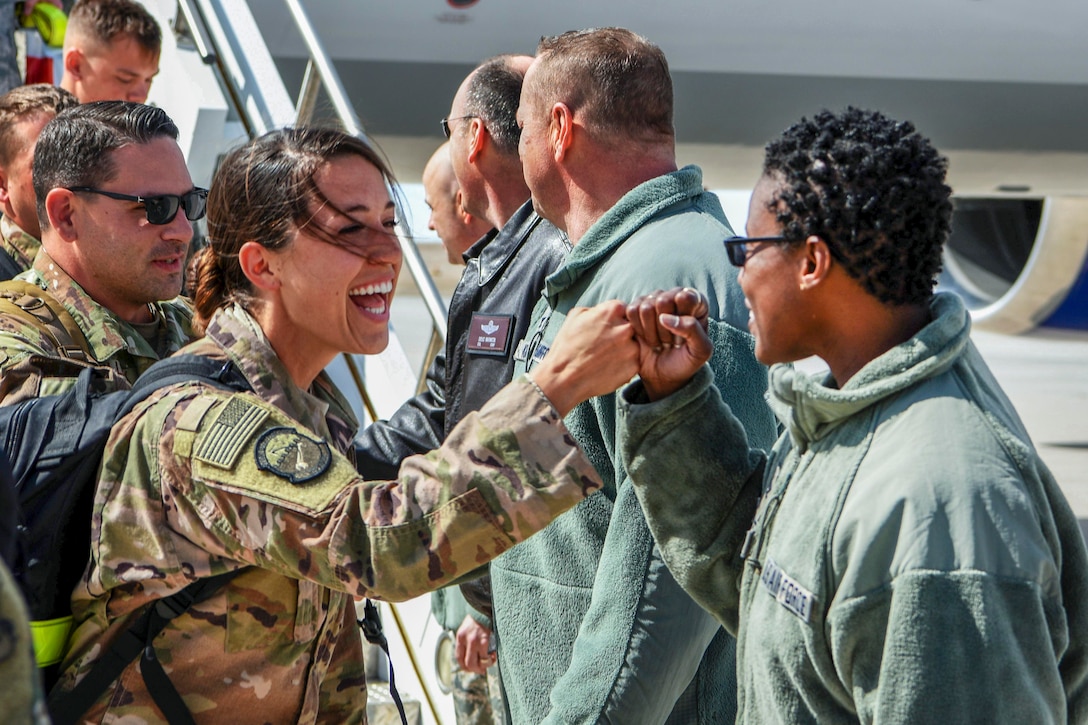 An airman smiles and fist-bumps a fellow airman, as other troops linger in the background by the stairs of an aircraft.