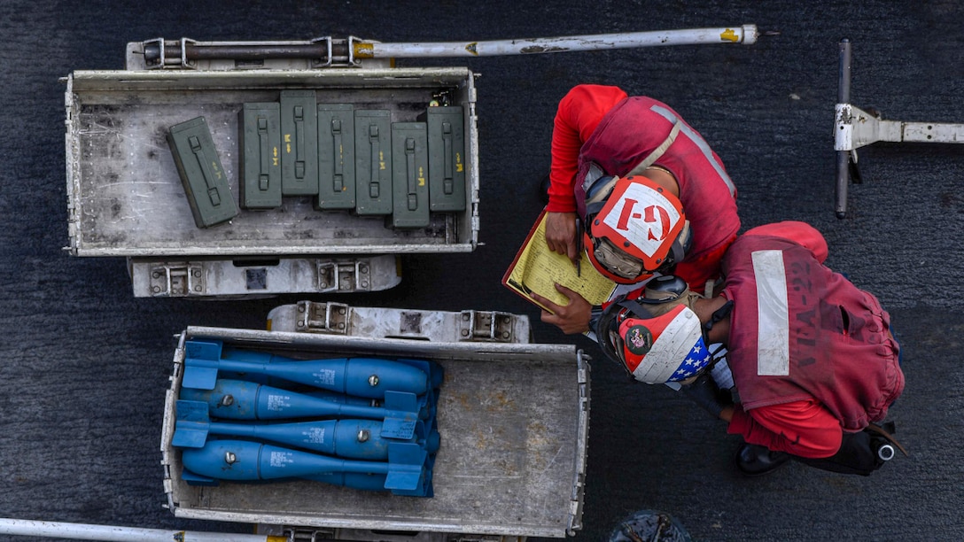 Sailors in red, seen from overhead, check a list while standing by two carts of munitions.