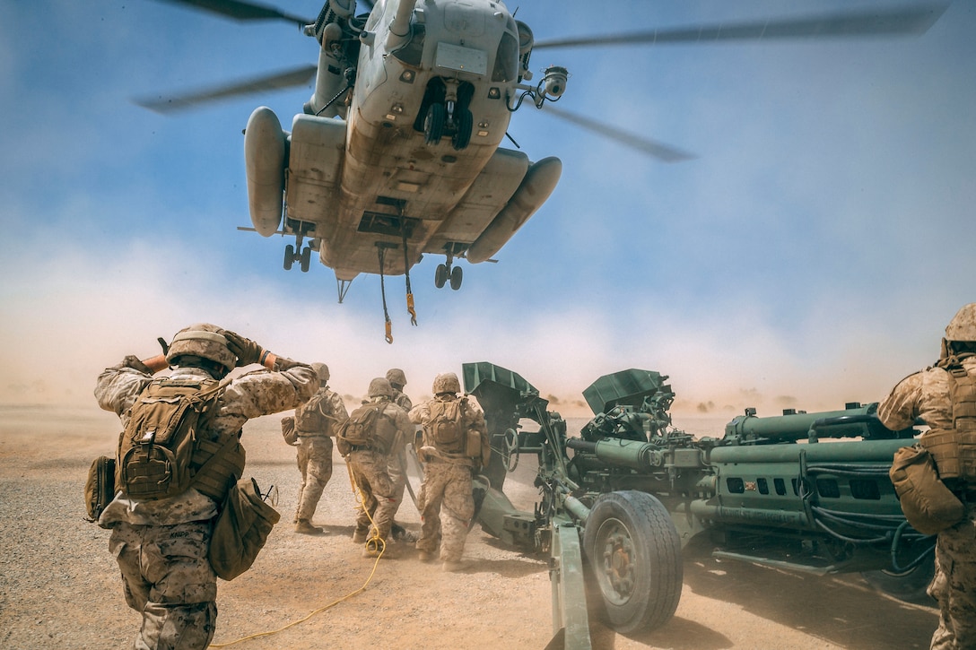 Marines brace themselves underneath a hovering helicopter, which kicks up dust, as a howitzer sits nearby.