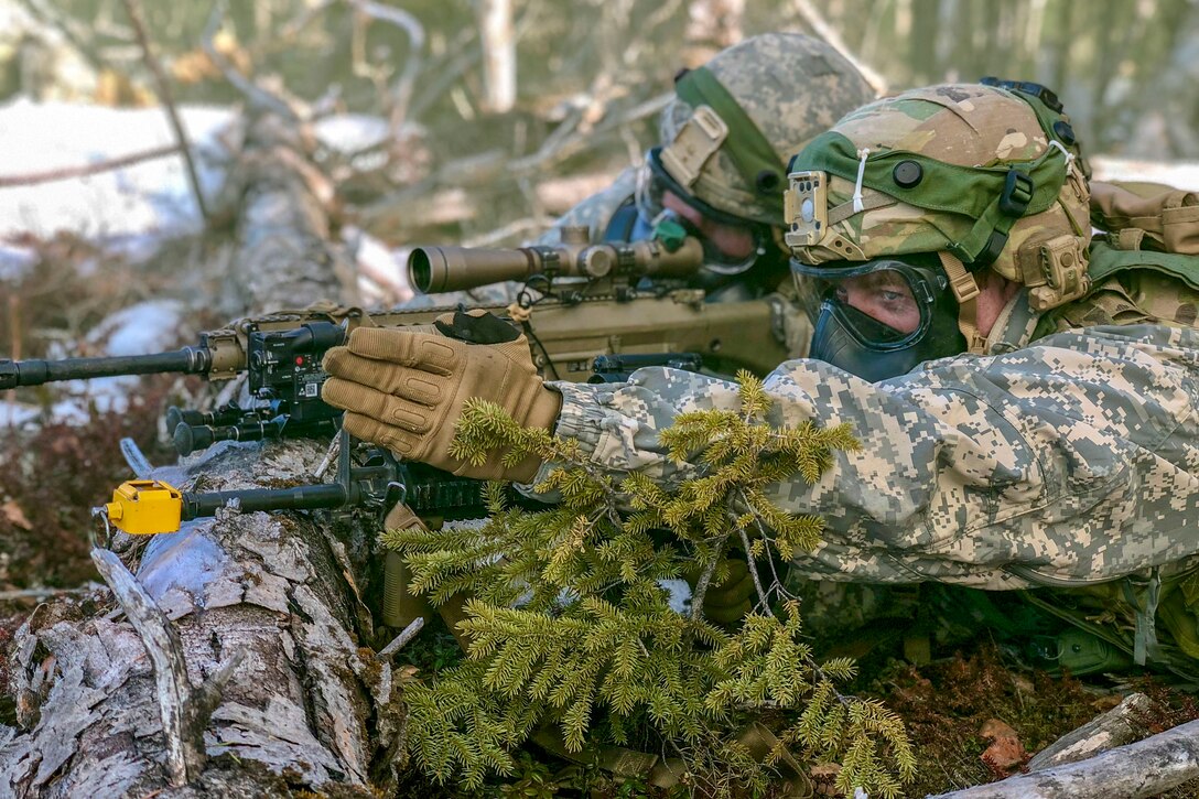 Two soldiers wearing gas masks lie prone in a wooded area and aim a weapon propped on a log.