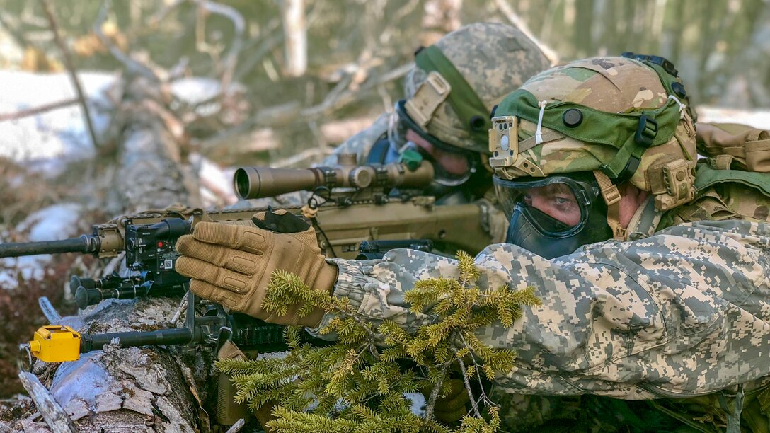 Two soldiers wearing gas masks lie prone in a wooded area and aim a weapon propped on a log.