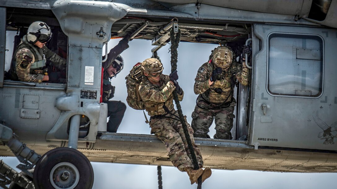 A service member holds onto a rope just outside the open door of a hovering helicopter.