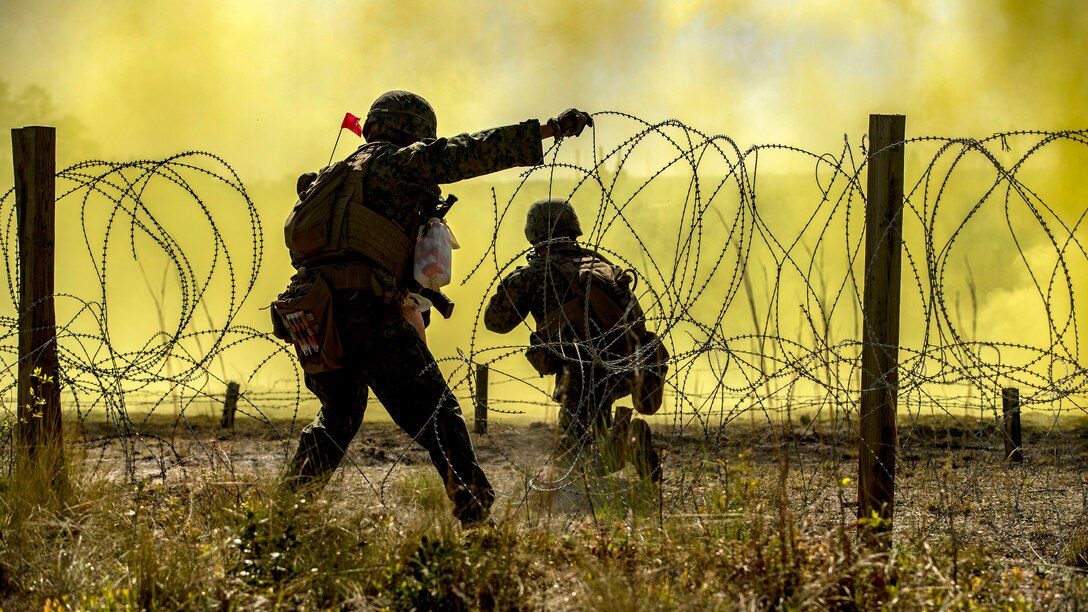 A Marine holds a breached section of barbed wire as another kneels on the other side of it, framed by yellow smoke.
