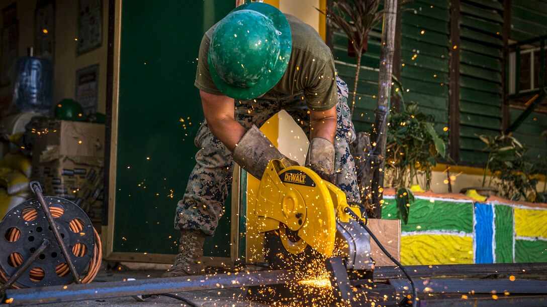 Sparks fly as a Marine in a green hard hat saws metal outside a school building