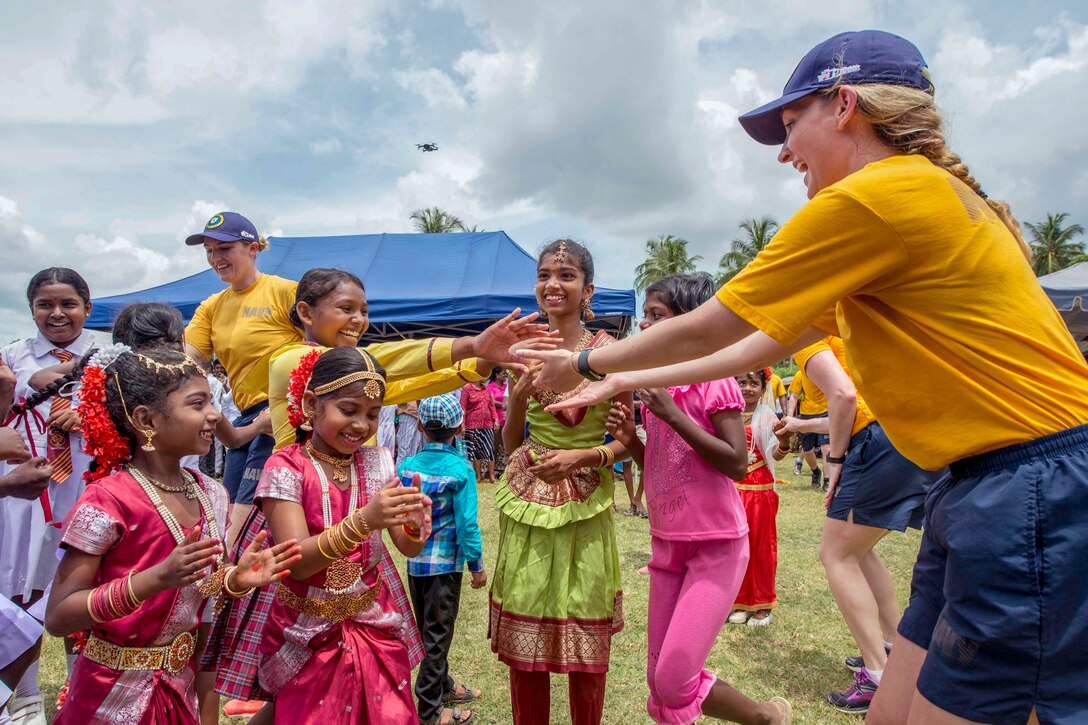 A smiling sailor extends her arms toward a smiling girl, who does the same amid a small crowd of other sailors and girls.