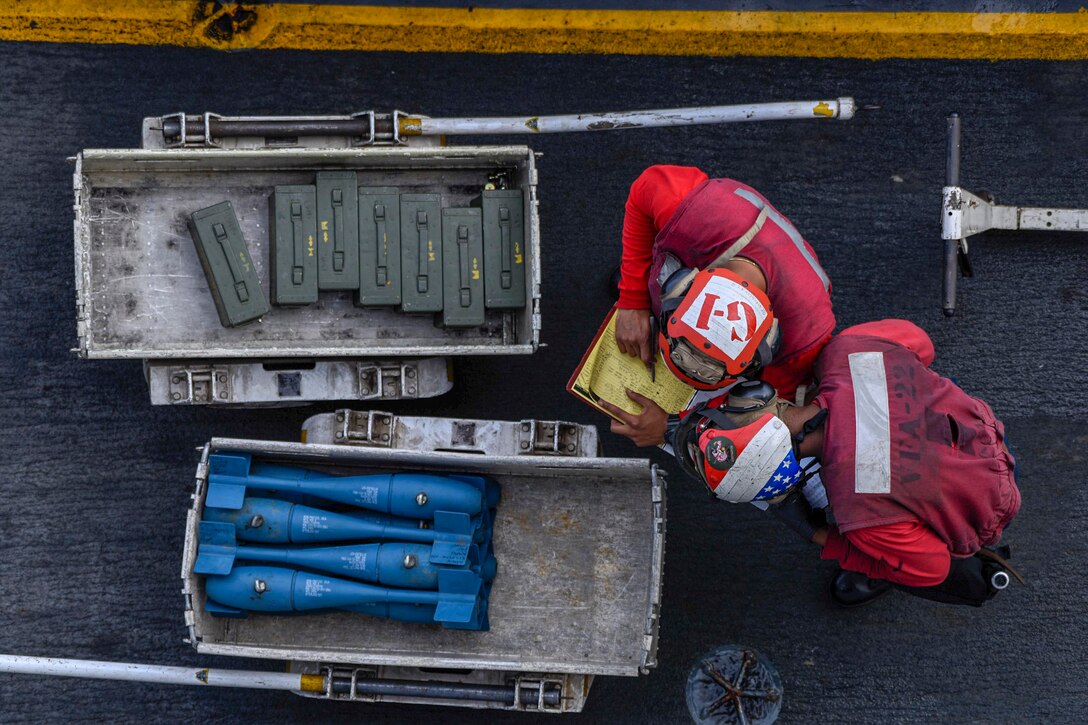 Sailors in red, seen from overhead, check a list while standing by two carts of munitions.