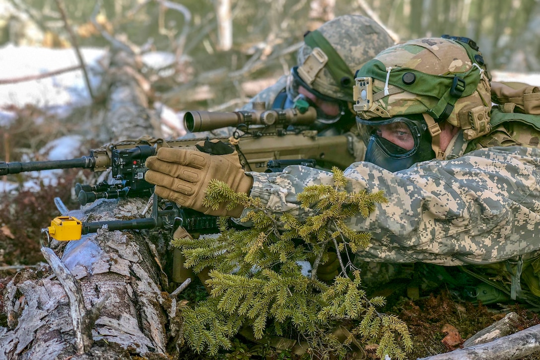 Two soldiers wearing gas masks lie prone in a wooded area and aim a weapon propped on a log.