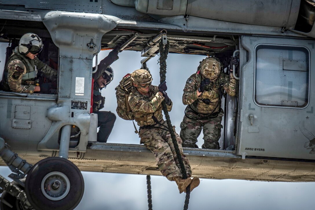 A service member holds onto a rope just outside the open door of a hovering helicopter.