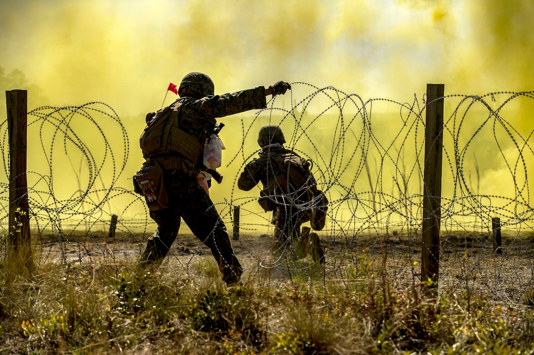 A Marine holds a breached section of barbed wire as another kneels on the other side of it, framed by yellow smoke.