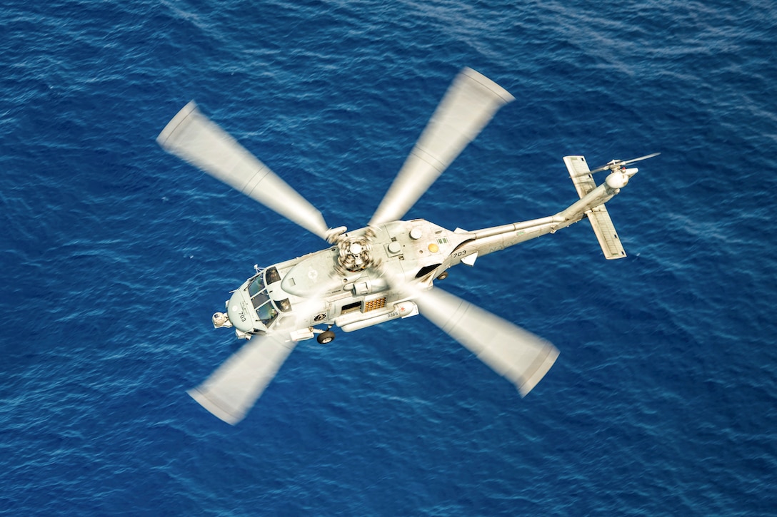 A helicopter, seen from overhead, flies over blue water.