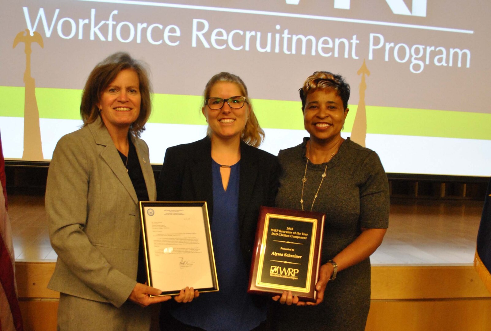 Three women stand together holding an award and a letter honoring one of them.