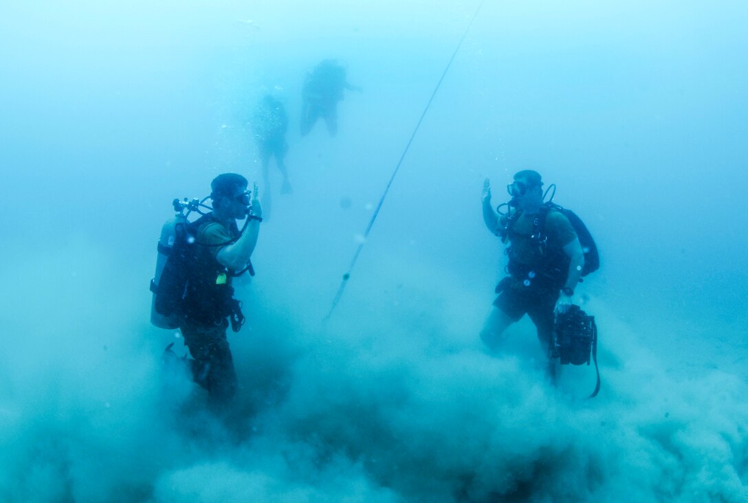 Two divers stand facing each other under water, each raising one hand.