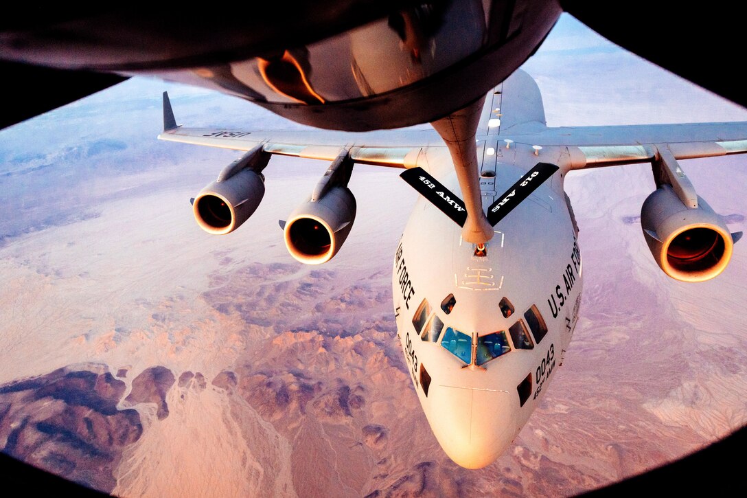 An aircraft receives fuel from the boom of another aircraft over pink desert terrain.