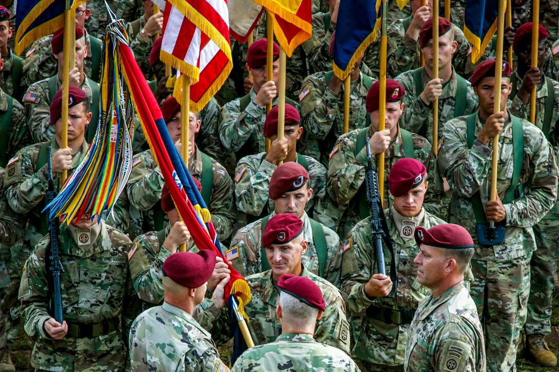 A soldier passe a guidon to another soldiers as others stand in formation with flags behind them.