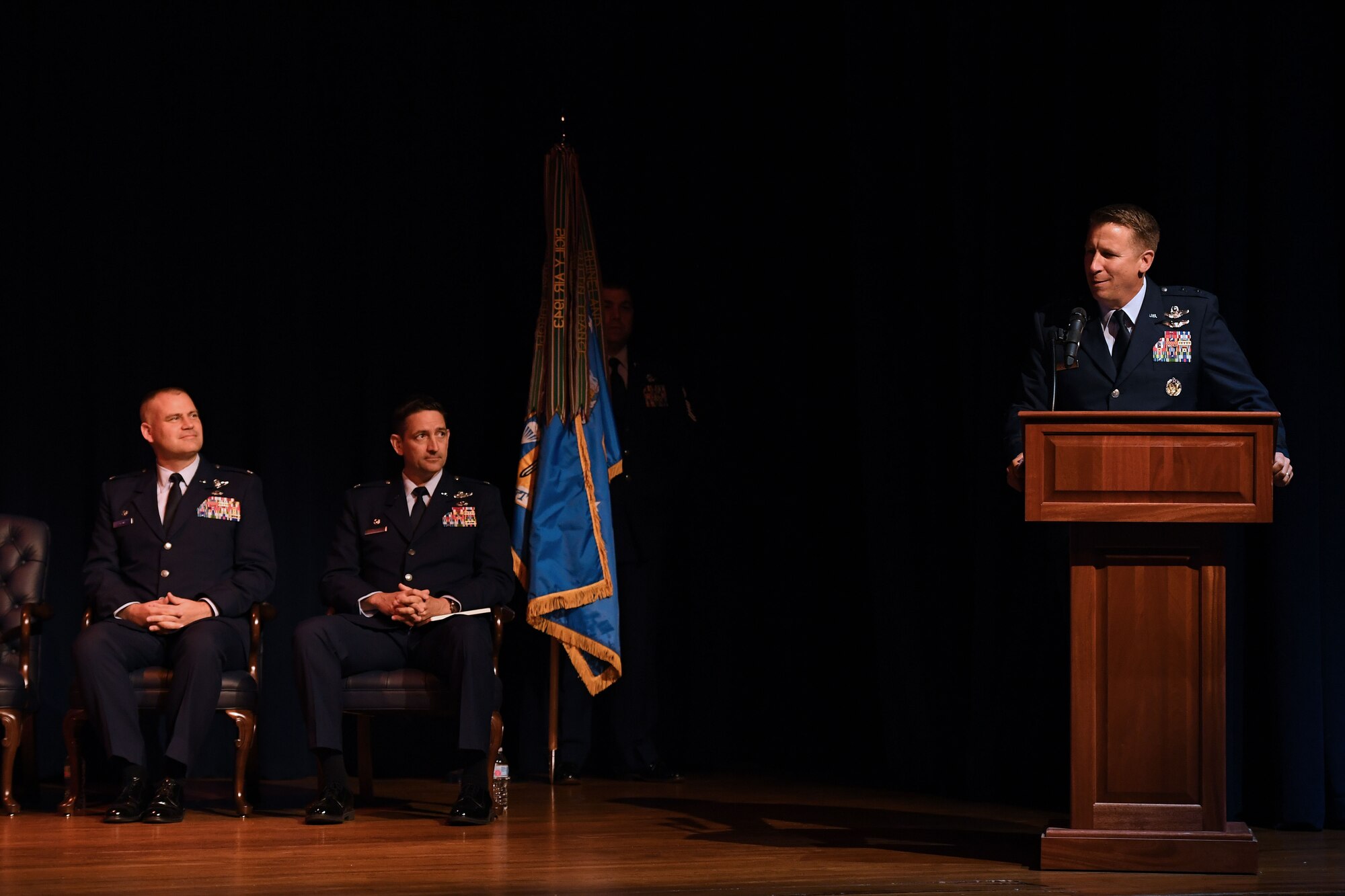 Two men sit on left side of screen, while one man talks at a podium on right side of screen.