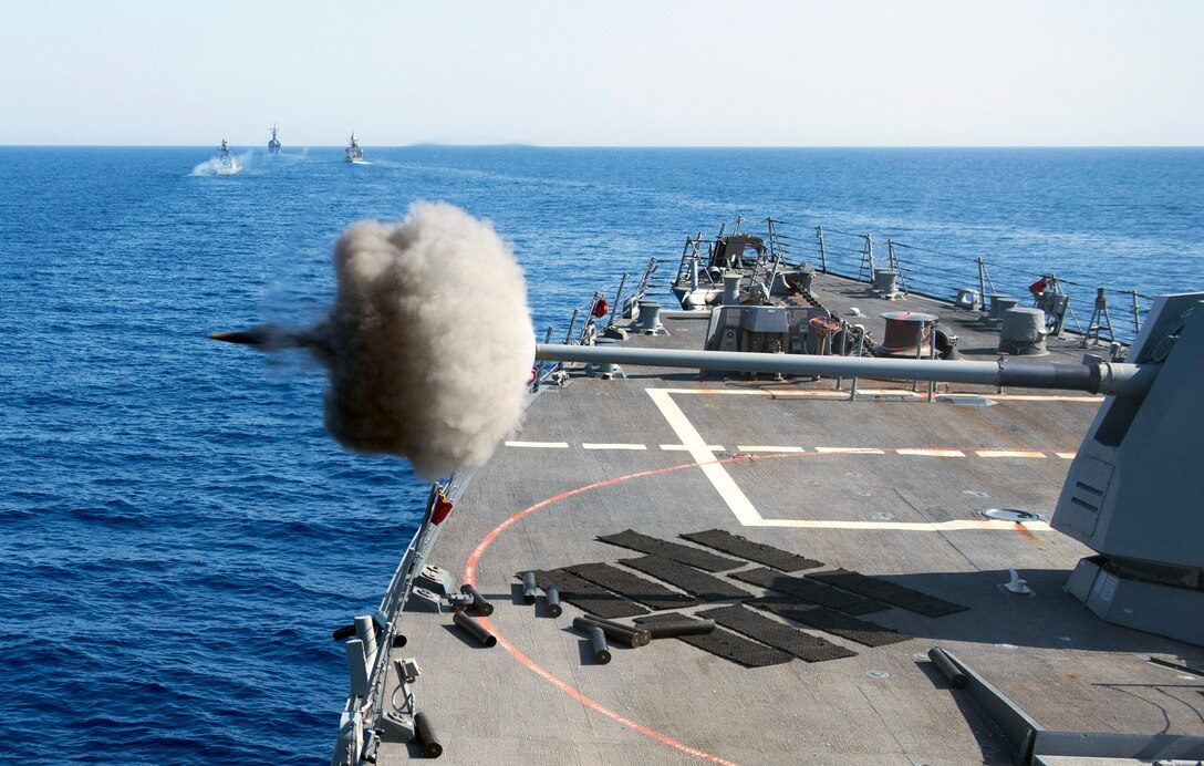 A projectile and burst of gray smoke shoots out from a gun on a ship in blue waters.