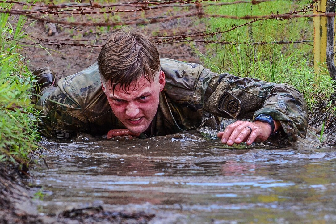 A soldier low crawls through water under the wire obstacle.