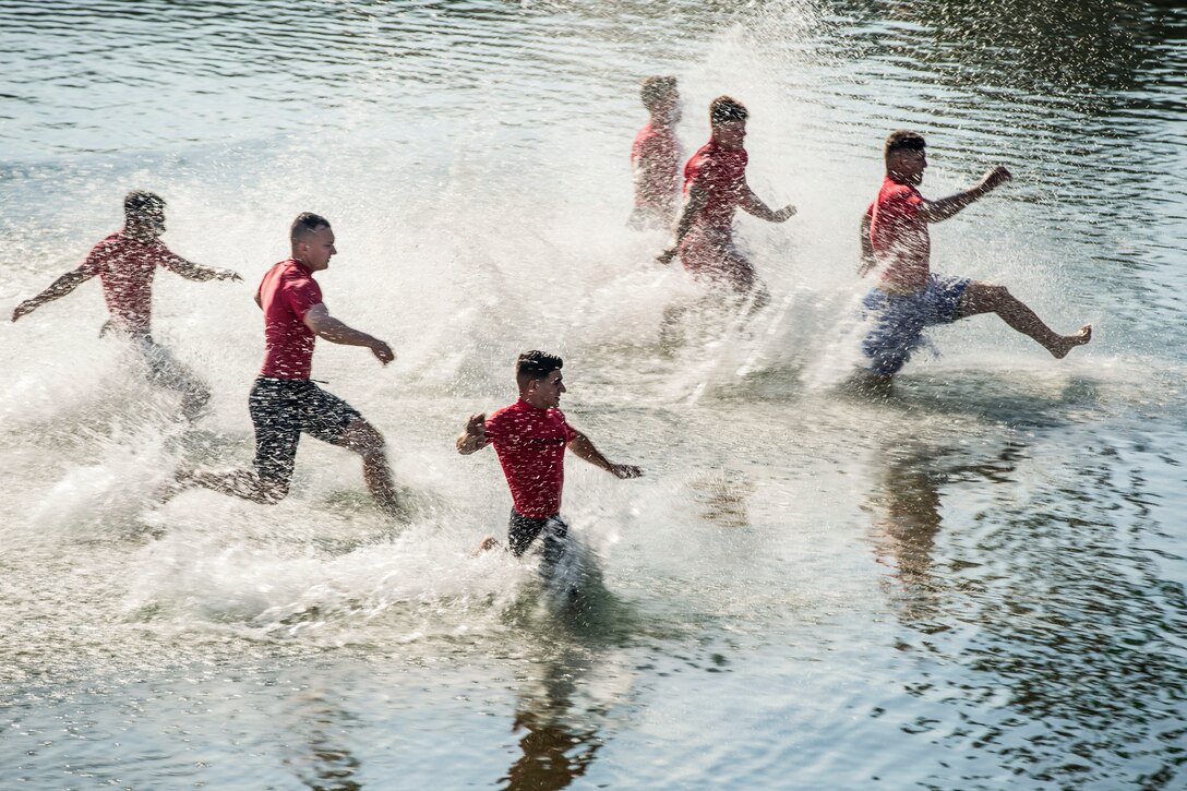 Marines in red shirts and shorts running through knee-deep water.