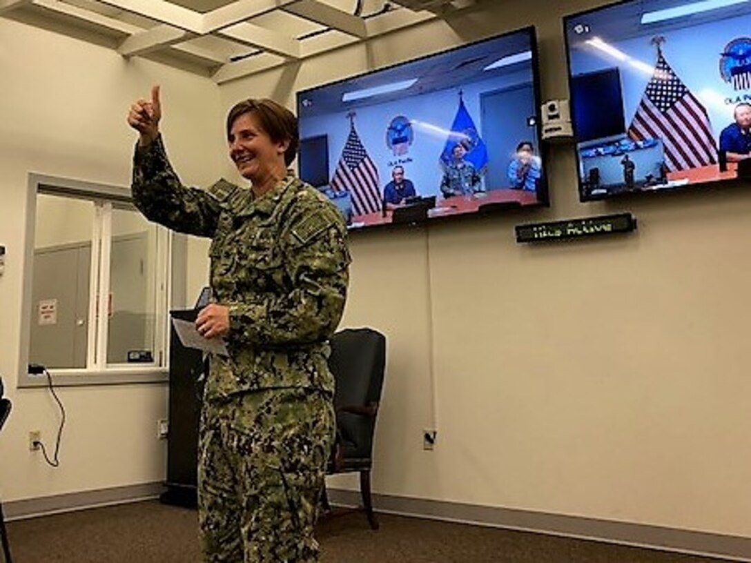 Navy captain in fatigues, addressing audience, holdling 'thumbs up' gesture