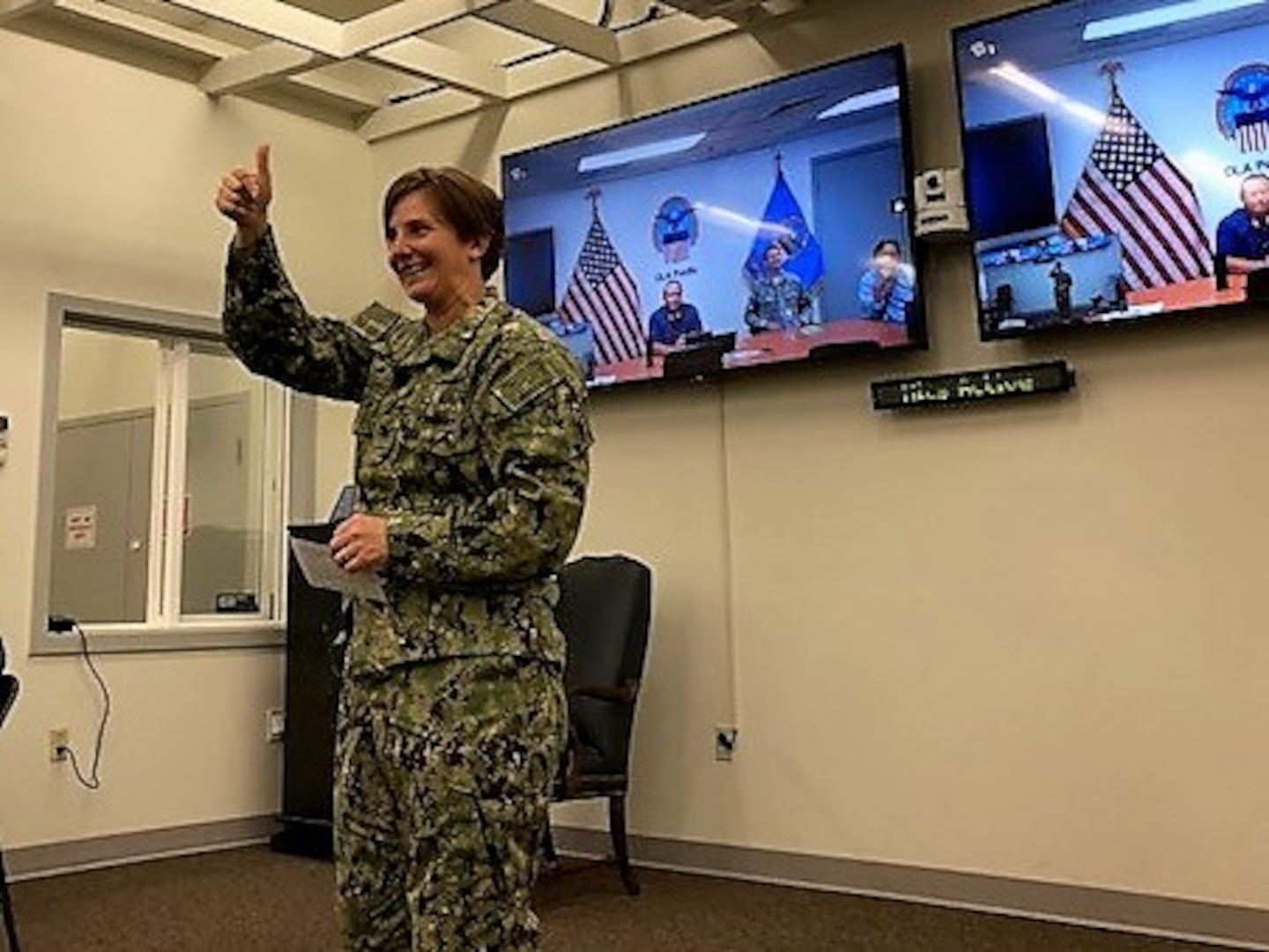 Navy captain in fatigues, addressing audience, holdling 'thumbs up' gesture