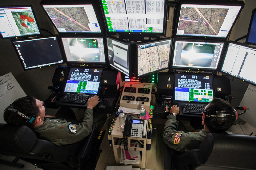 Two Air National Guard pilots sit in front of a wall of computer screens  while flying a remotely piloted aircraft.