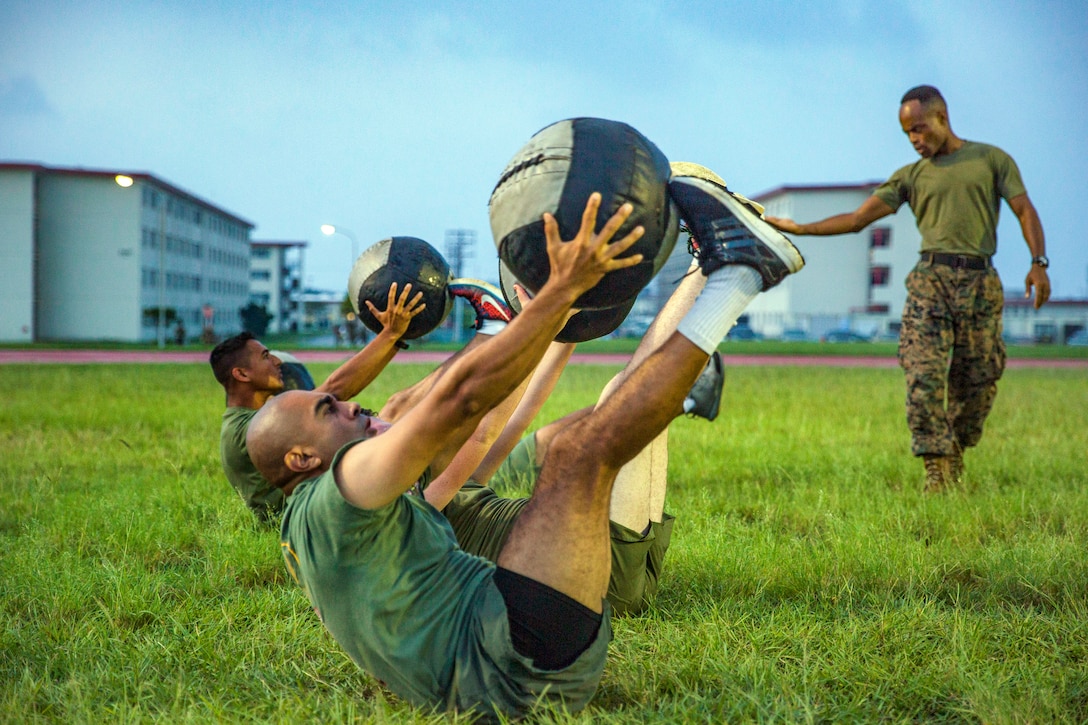 A line of Marines lay on the ground with medicine balls while another Marine stands and watches.