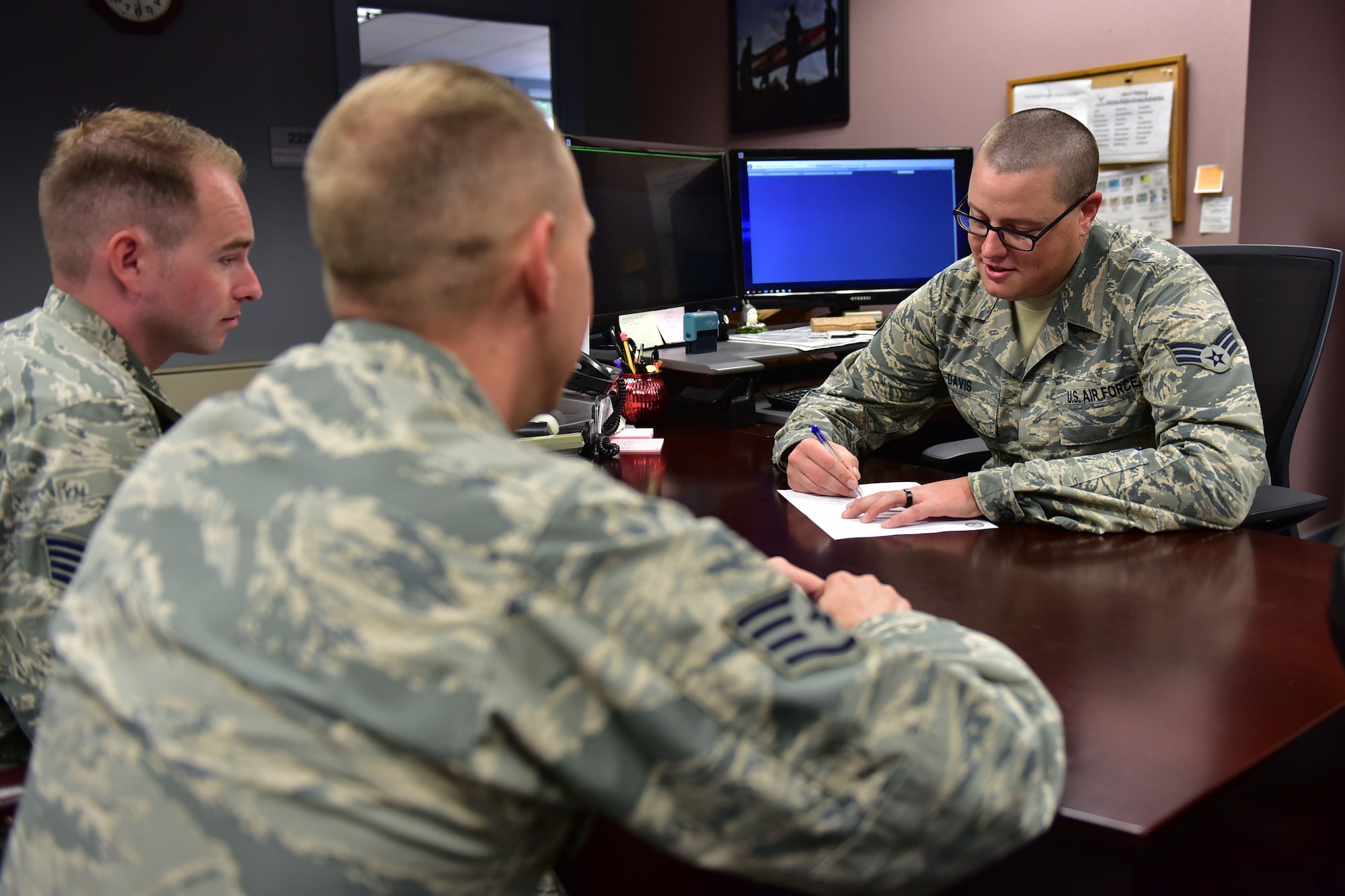 One man sits at a desk and signs a piece of paper, while two men sit on the other side of the desk and watch.