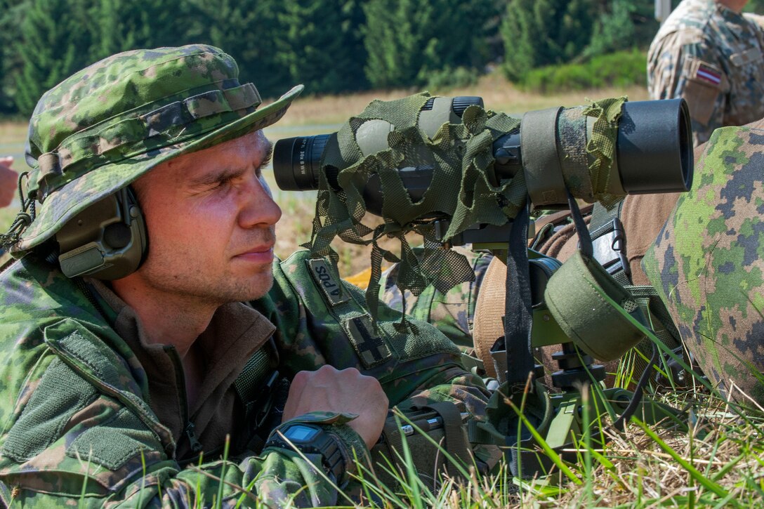 A Finland soldier uses a spotting scope to watch targets being hit by rounds fired by a sniper.