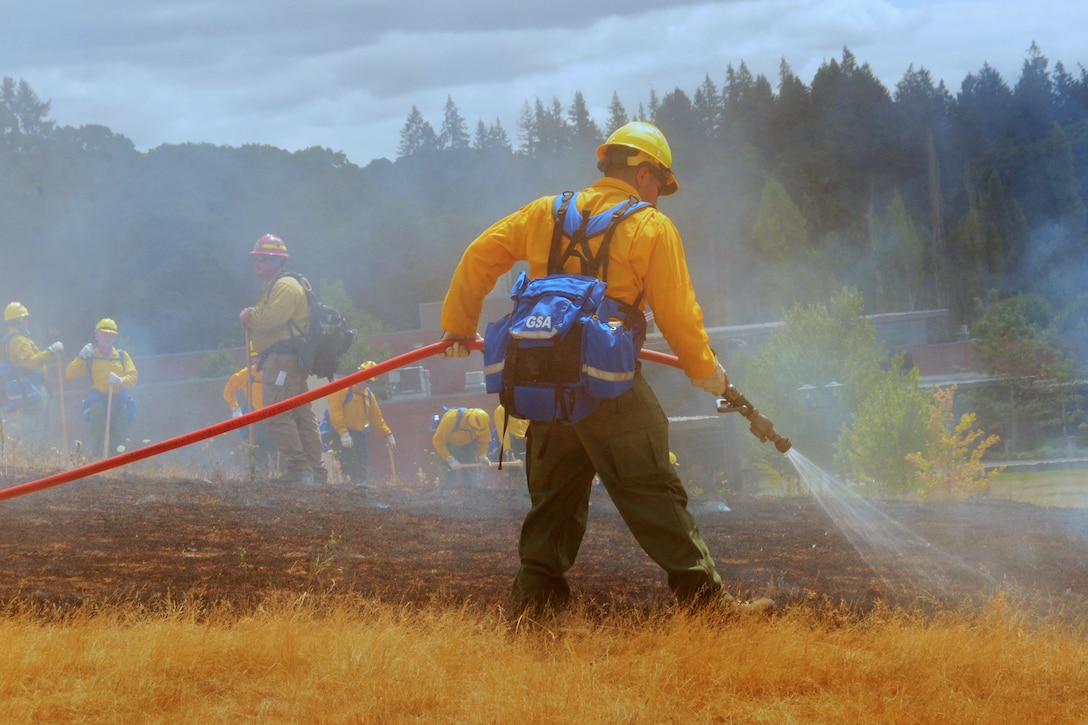 A soldier sprays water from a fire hose onto to smoldering grass.