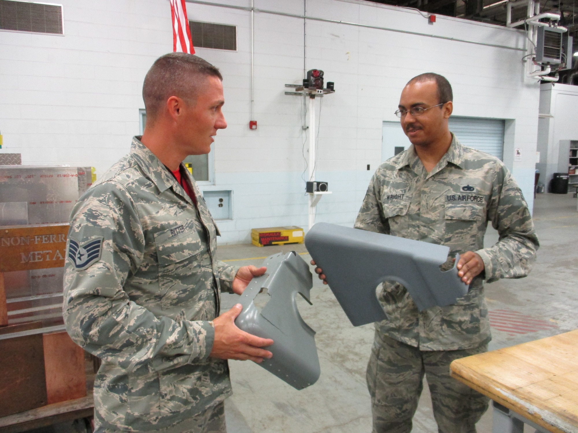 Staff Sgt. Christopher Ritter, 513th Maintenance Squadron Section Chief, and Staff Sgt. Michael Wright, 552nd Maintenance Squadron Aircraft Metals Technology, discuss the differences 3-D printing bring to the AWACS fabrications shop.  The damaged part has a two-year backlog for replacement parts and requires riveted reinforcement, while the printed part has reinforcements belt in  and can be printed in a single day. (U.S. Air Force photo by Master Sgt. Andrew Stephens)