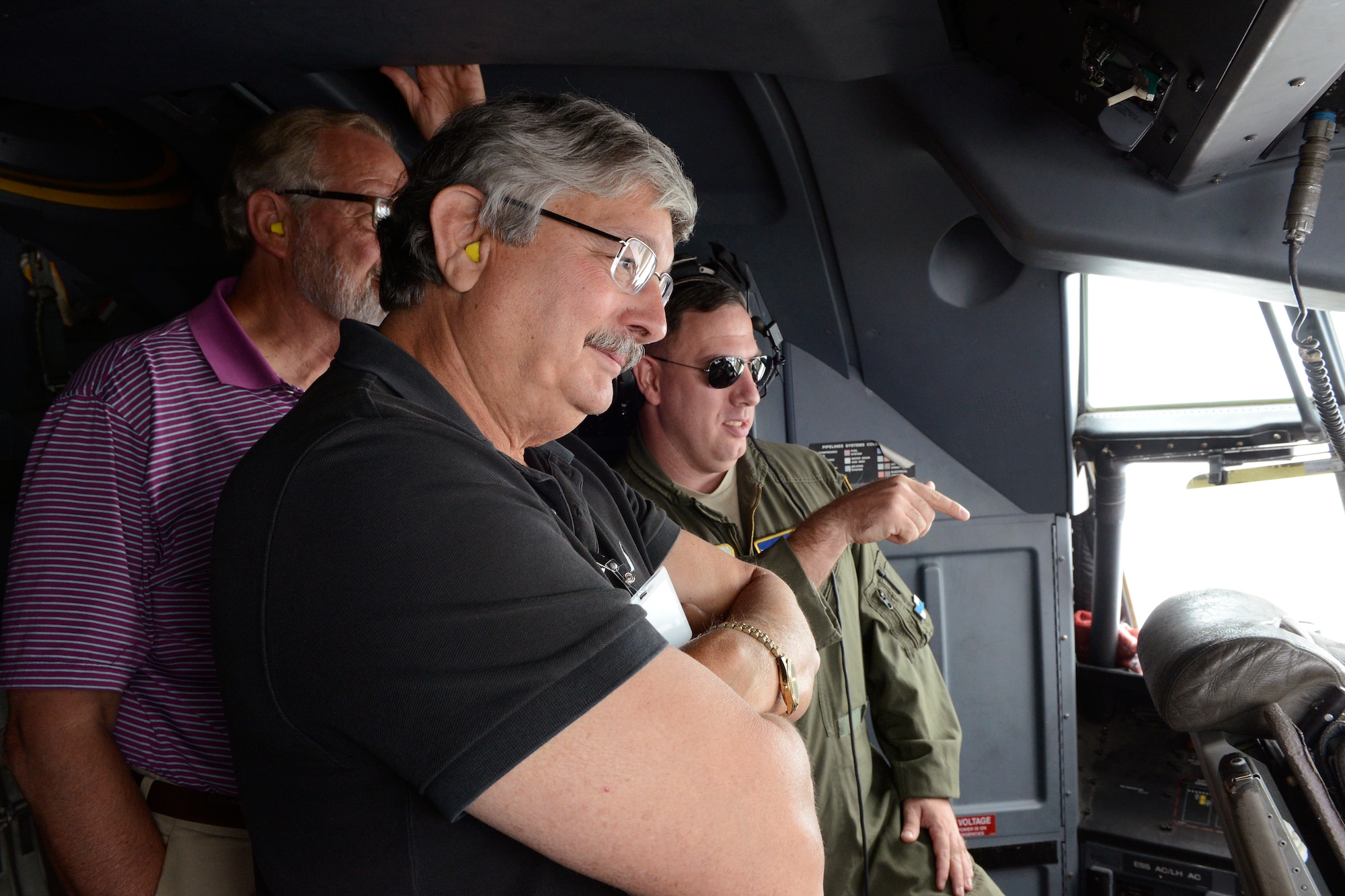 Two white male civilians listen to a white male in a green flight suit in the cockpit of a C130J airplane.