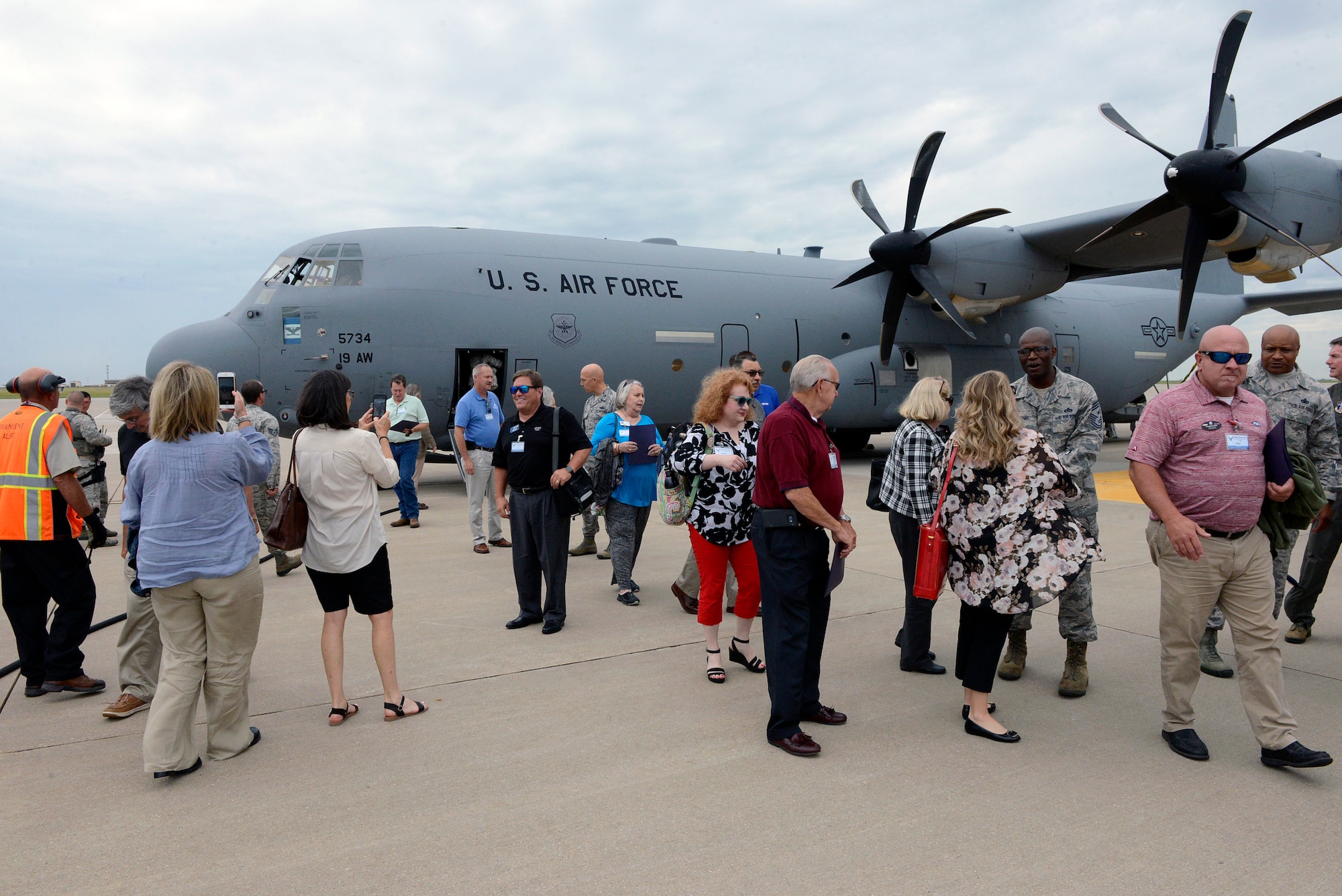 Approximately twenty people gather on the concrete of the flight line after departing a grey C-130J Air Force airplane in the background.