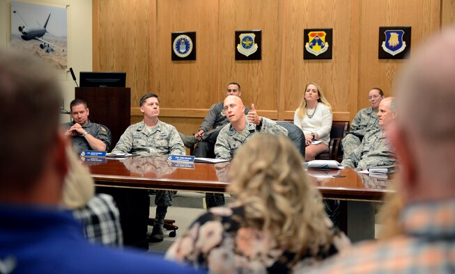 Four uniformed white males sit at a brown conference table to brief a group of civilians sitting in front of them.