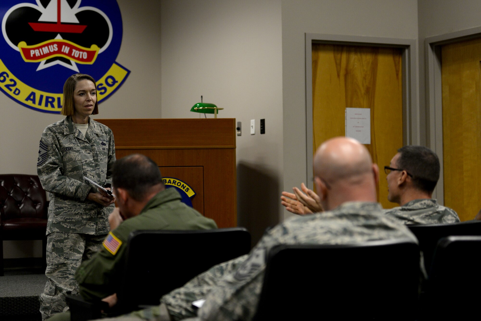A woman in the Airman Battle Uniform looks at a man speaking to her as she stands in front of a room full seated individuals wearing ABUs.
