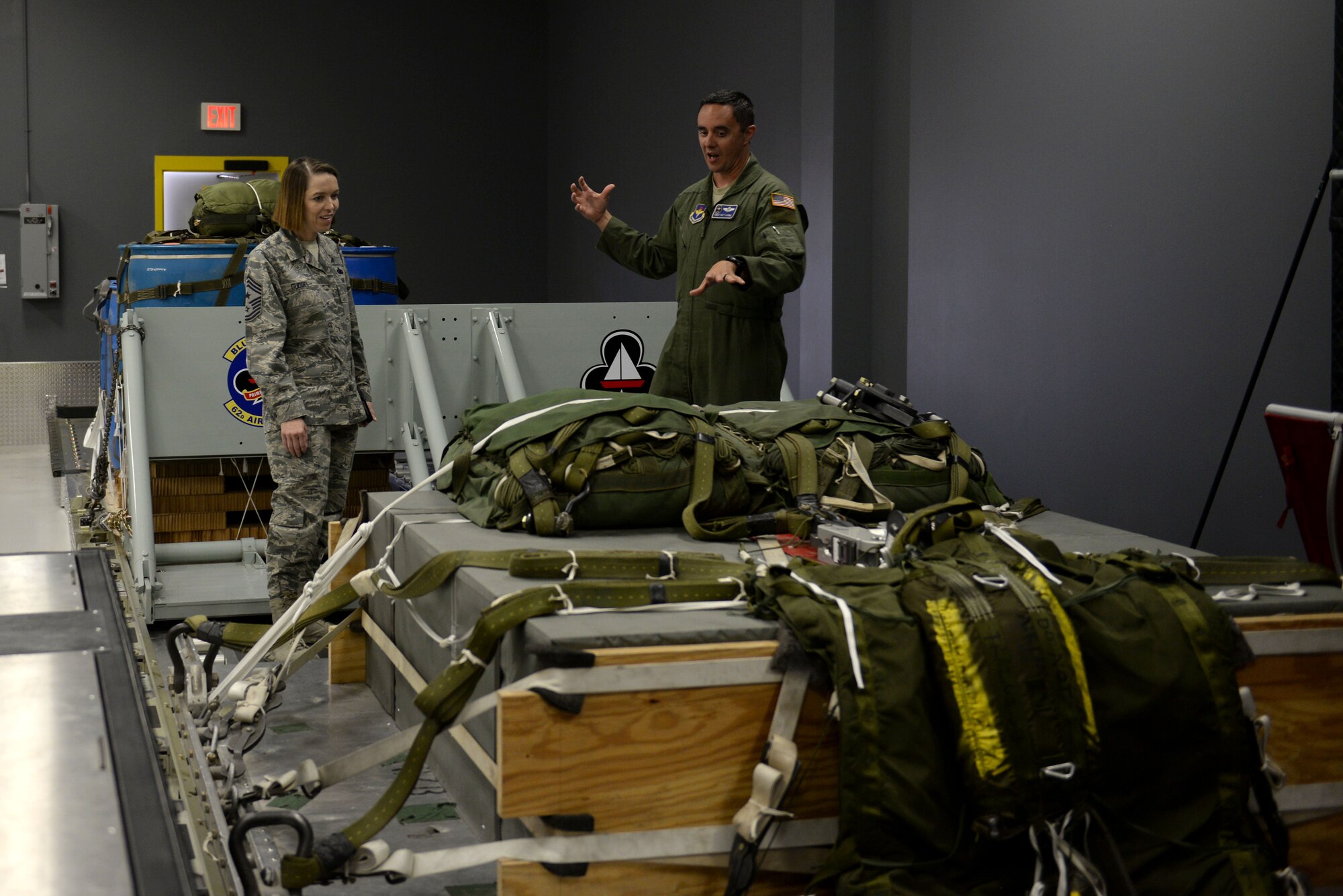 A man wearing the Air Force flight suit motions toward heavy equipment as a woman in the Airman Batte Uniform looks at the heavy equipment.