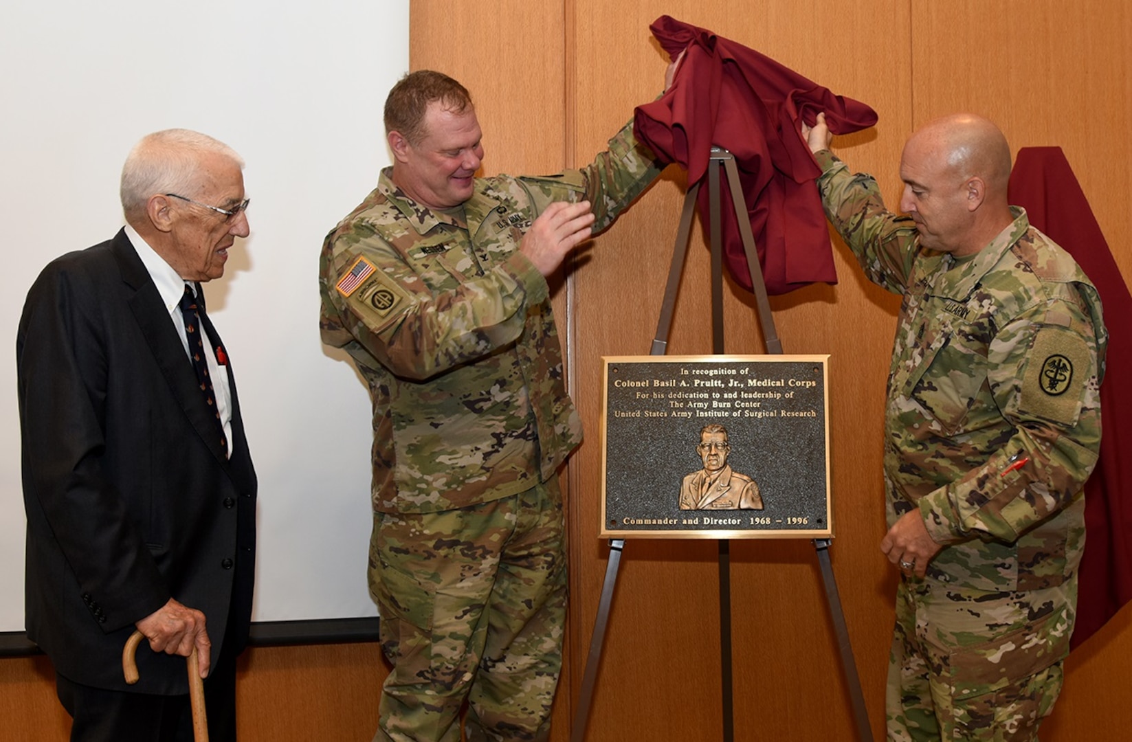Col. (Dr.) Shawn C. Nessen (center), U.S. Army Institute of Surgical Research commander, and the USAISR Senior Enlisted Advisor, Sgt. Maj. William “Dave” Poist Jr., unveil a bronze plaque for Dr. Basil Pruitt Jr., during the USAISR 70thAnniversary Symposium July 18 at Joint Base San Antonio-Fort Sam Houston.