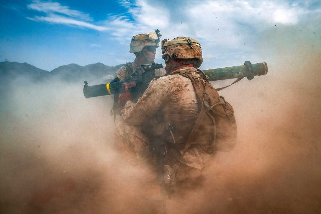 Two marines stand in a dusty wind carrying a shoulder-mounted weapon.
