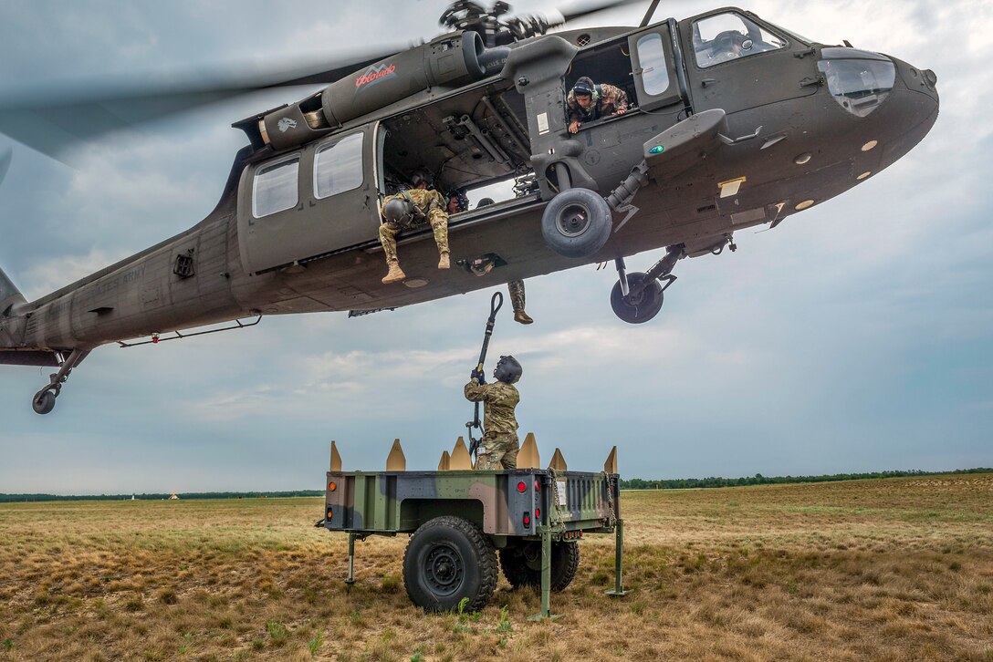 A soldier attaches a hook to the bottom of a helicopter hovering overhead.