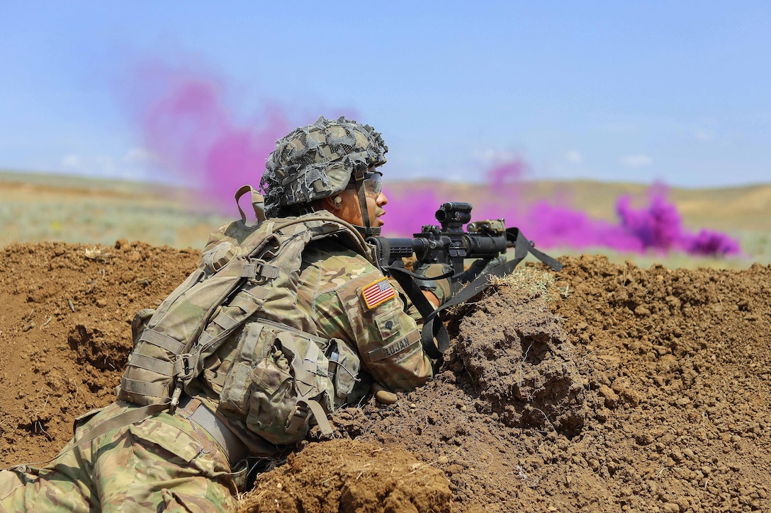 A soldier takes cover behind a berm as purple smoke blows in front of him.