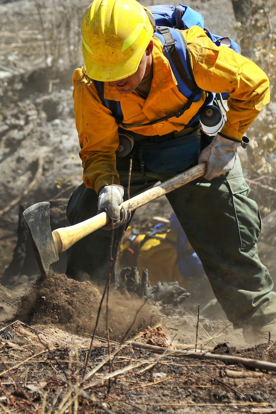 An Oregon Army National Guardsman uses a pickaxe to dig up charred dirt and brush while fighting a wildfire.