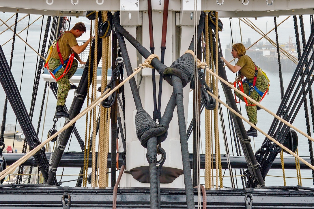 Sailors climb the main mast of a ship.