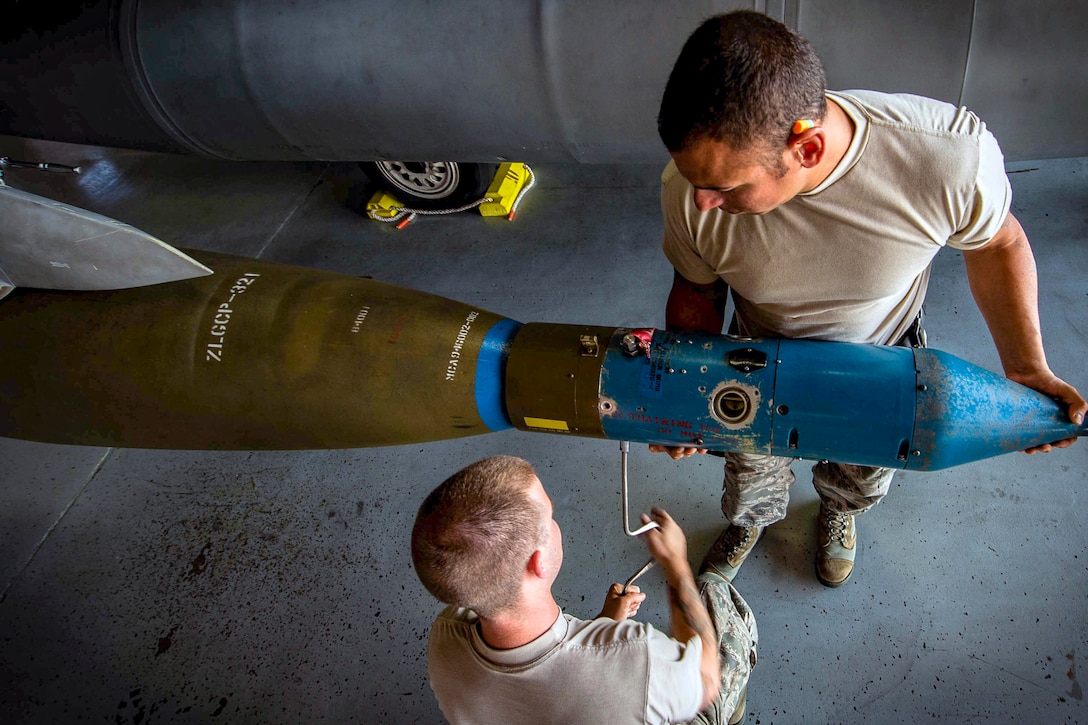 Two airmen hold large bomb while removing equipment from around it.