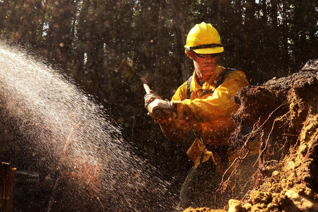 An airman uses a pickaxe to clear brush along a fire line.