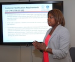 Woman speaking while standing in front of an on screen presentation.