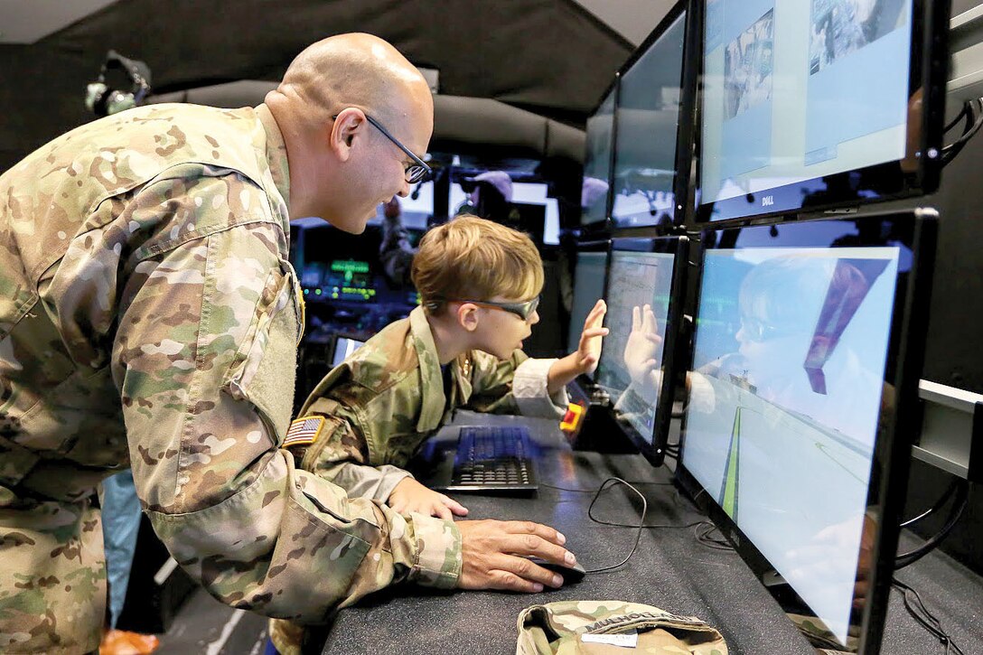 A soldier and a young boy stand in front of computer monitors.