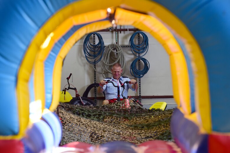 Col. Larry Shaw rides a tricycle toward the low-crawl portion of the obstacle course during a combat dining in at Grissom Air Reserve Base, Ind., Aug. 18, 2018. Grissom held its first combat dining in since 2007, where Airmen faced several fun challenges and were encouraged to show off their homemade uniforms. (U.S. Air Force Photo / Staff Sgt. Christopher Massey)