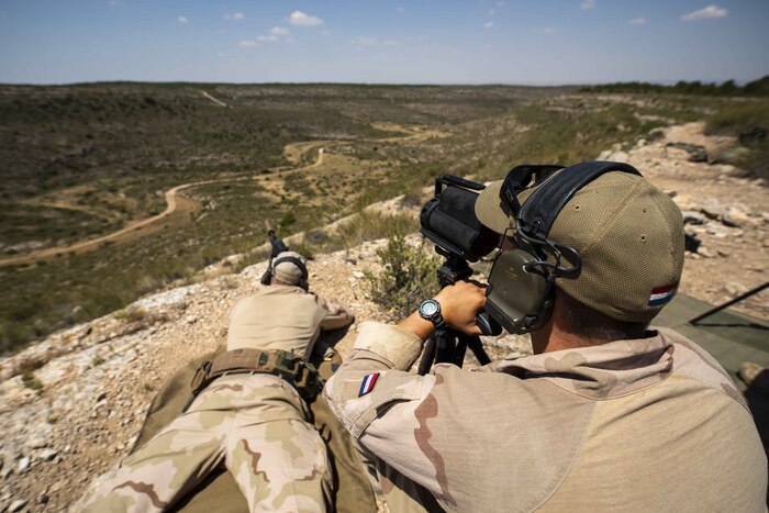 One service members lies on the ground firing a rifle while another sits behind him.